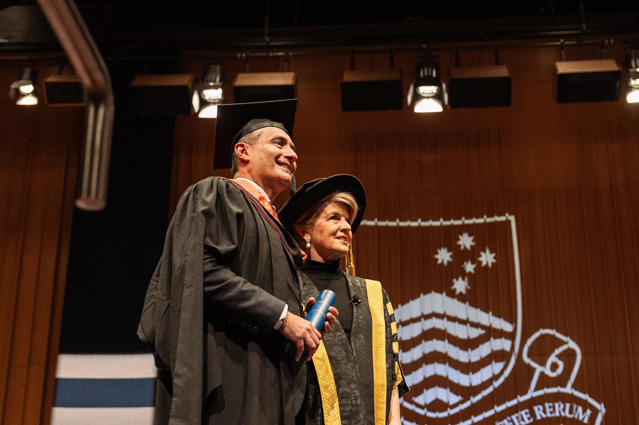 Steven Kourras standing on stage with Julie Bishop in graduation robes, holding his Testamur